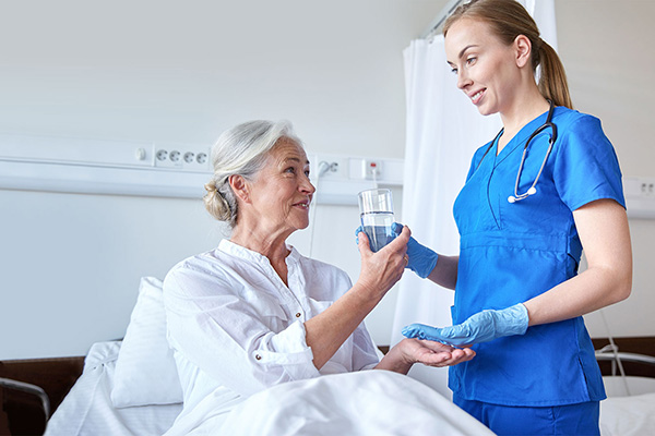 An elderly woman in bed and a caregiver standing with medication and a glass of water